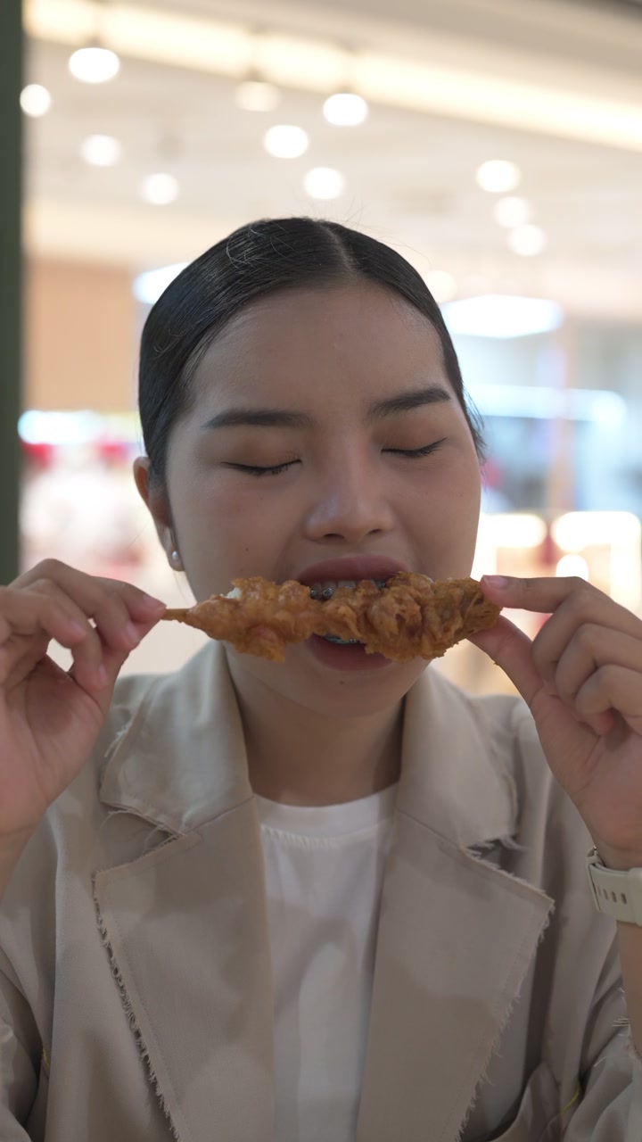 Close-up seorang pengusaha wanita muda Thai Isan yang lapar sedang makan sate ayam goreng di meja pujasera.