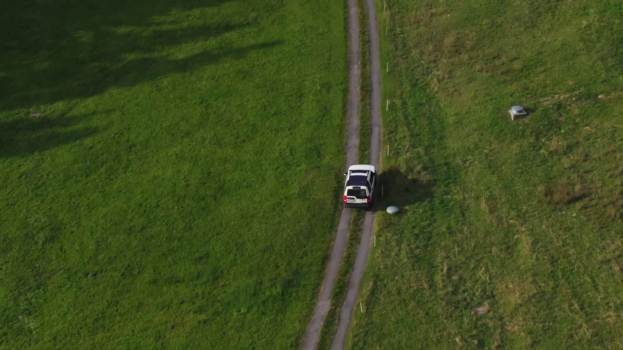 Drone view of a white car driving along a small dirt road through green fields on a sunny day. Minimalistic countryside landscape symbolizing travel, freedom, and exploration