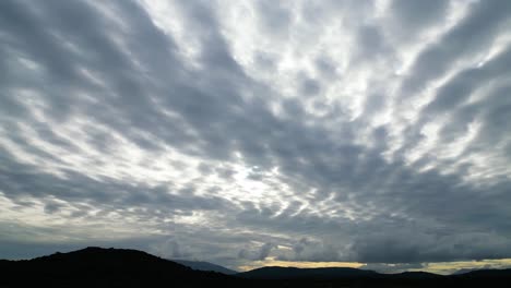 aerial shot with camera pan taking the sunset sky full of high gray blue clouds and orange colors of the sunset over the silhouette of the mountains in blue and black