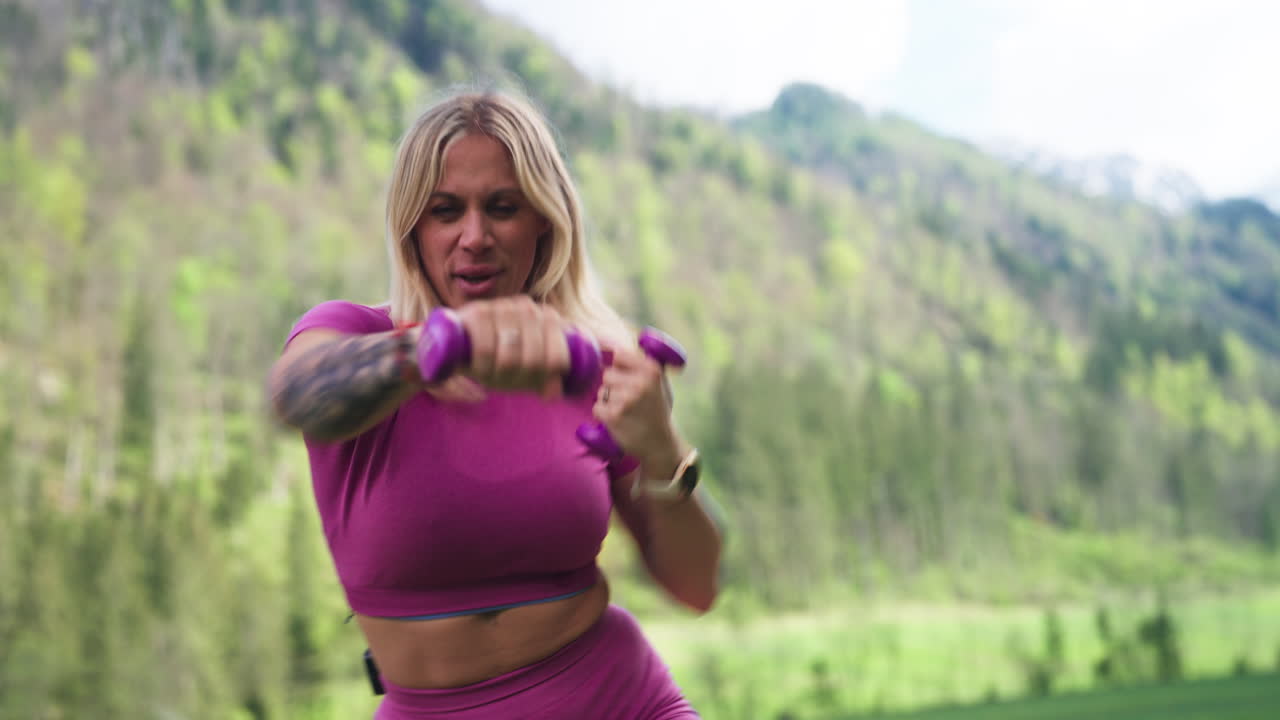 A Woman in a Pink Workout Outfit Exercises With Small Dumbbells in Logar Valley, Kamnik Alps, Slovenia - Handheld Shot