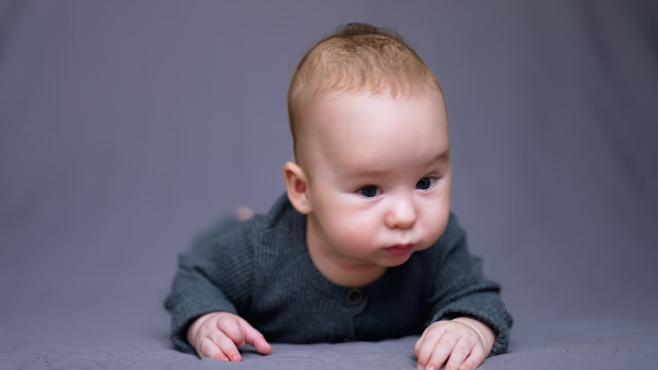 Adorable little child in grey clothes lies on belly in front of camera. Sweet funny boy with plump cheeks making faces. Close up.