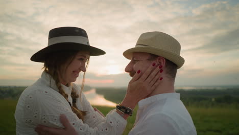 a wife in a white dress, wearing a hat and bracelets, smiles warmly as she lovingly touches her husband's face in a grassy field at sunset. he wears a white shirt and a hat