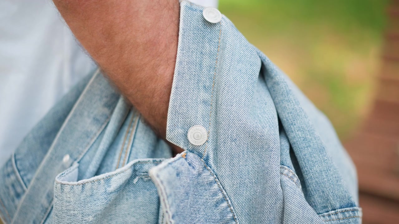 Primo piano di un uomo che tiene e sistema la sua giacca di jeans azzurra sul braccio, mettendo in mostra la trama del tessuto e i dettagli dei bottoni. La scena è ambientata all'aperto con una morbida illuminazione naturale e vegetazione.