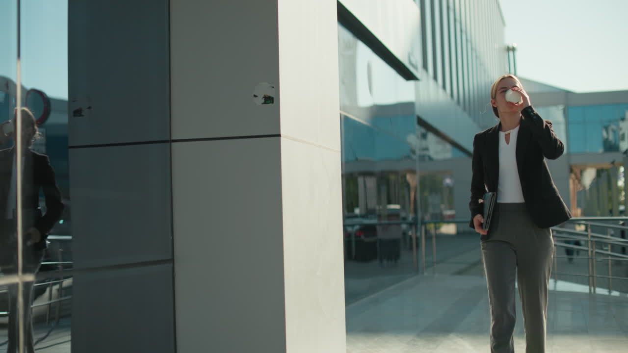 Lady in professional attire holding notepad walks confidently toward modern glass building while sipping drink, with sunlight casting her shadow on wall and cars parked in urban surroundings