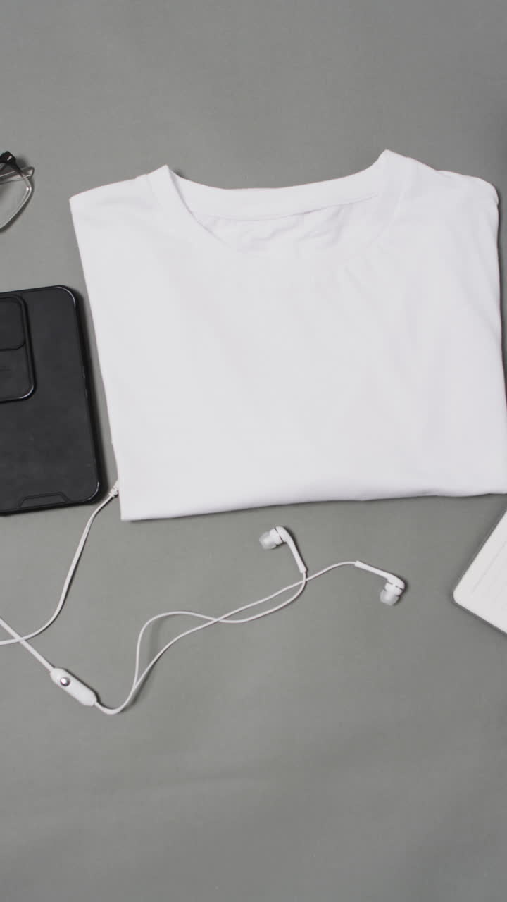 Vertical video of white t shirt, smartphone and earphones with copy space on grey background