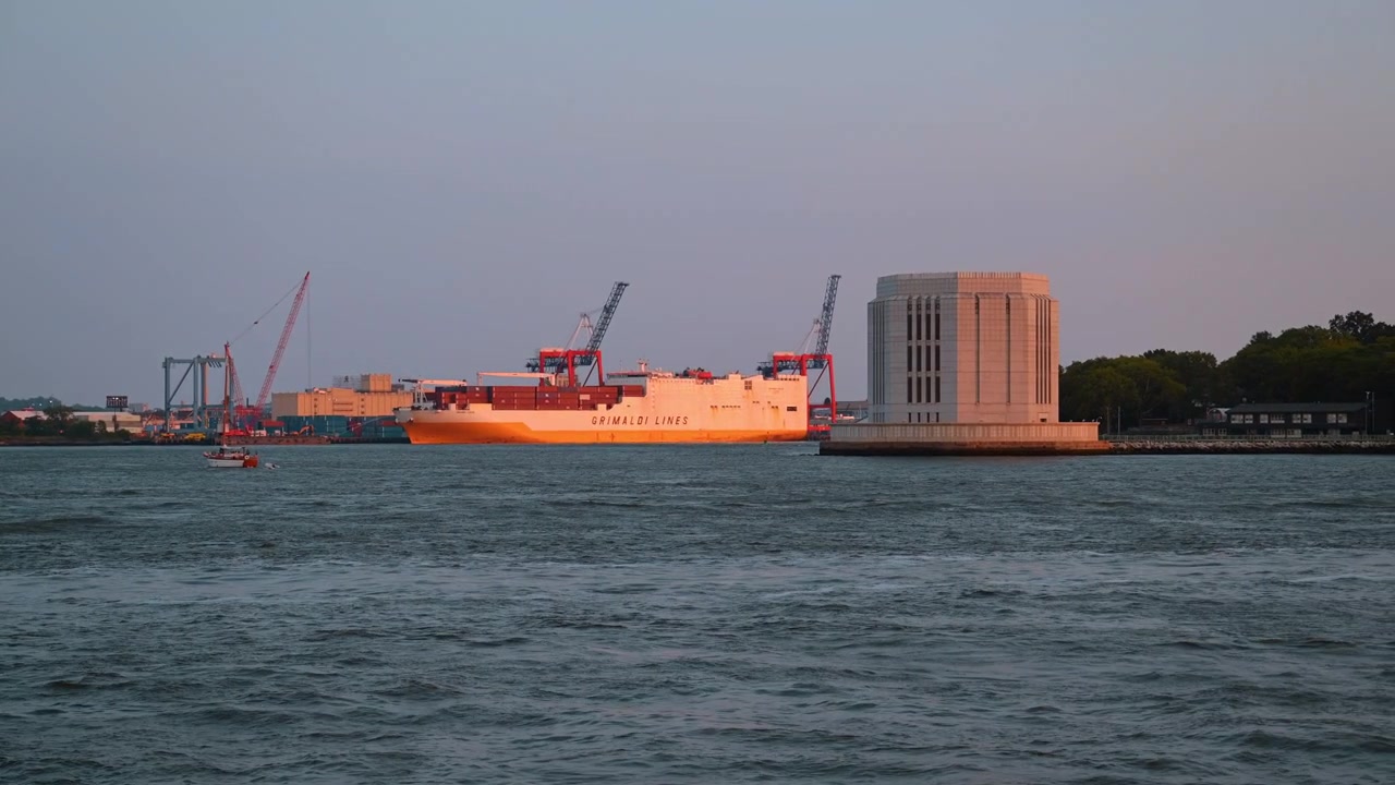 New York, USA, 25 May 2026: Cargo ship moving across water near ventilation tower in New York harbor. Orange and white commercial vessel floating past stone structure at evening light.