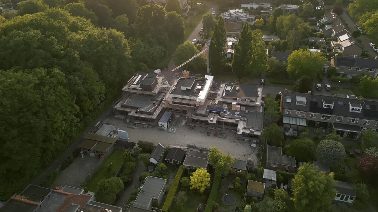 Aerial shot of a new housing construction project bordered by mature trees in a Dutch suburb, lit by evening sun.
