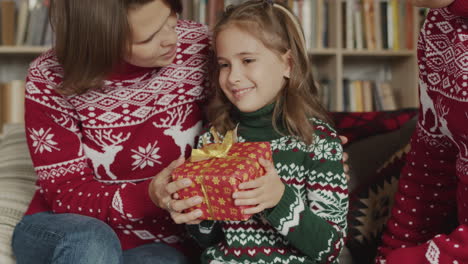 cute little girl holding and shaking christmas gift while sitting with her parents on sofa at home