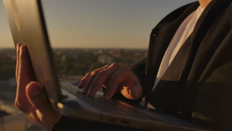 close-up: a programmer's hand typing on a laptop keyboard at sunset overlooking the roof. a businessman works remotely. freelancer performs work on vacation