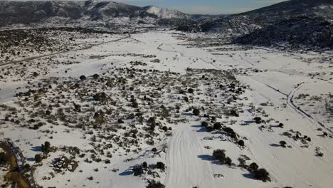 Vuelo-Del-Dron-A-La-Inversa-Centrándonos-En-Una-Llanura-Completamente-Nevada-Con-Arbustos-Y-Visualizamos-Un-Camino-De-Tierra-Despejado-De-Nieve-Para-Acceder-A-Fincas-Rurales-Con-Un-Fondo-De-Montañas-En-Invierno-ávila-España