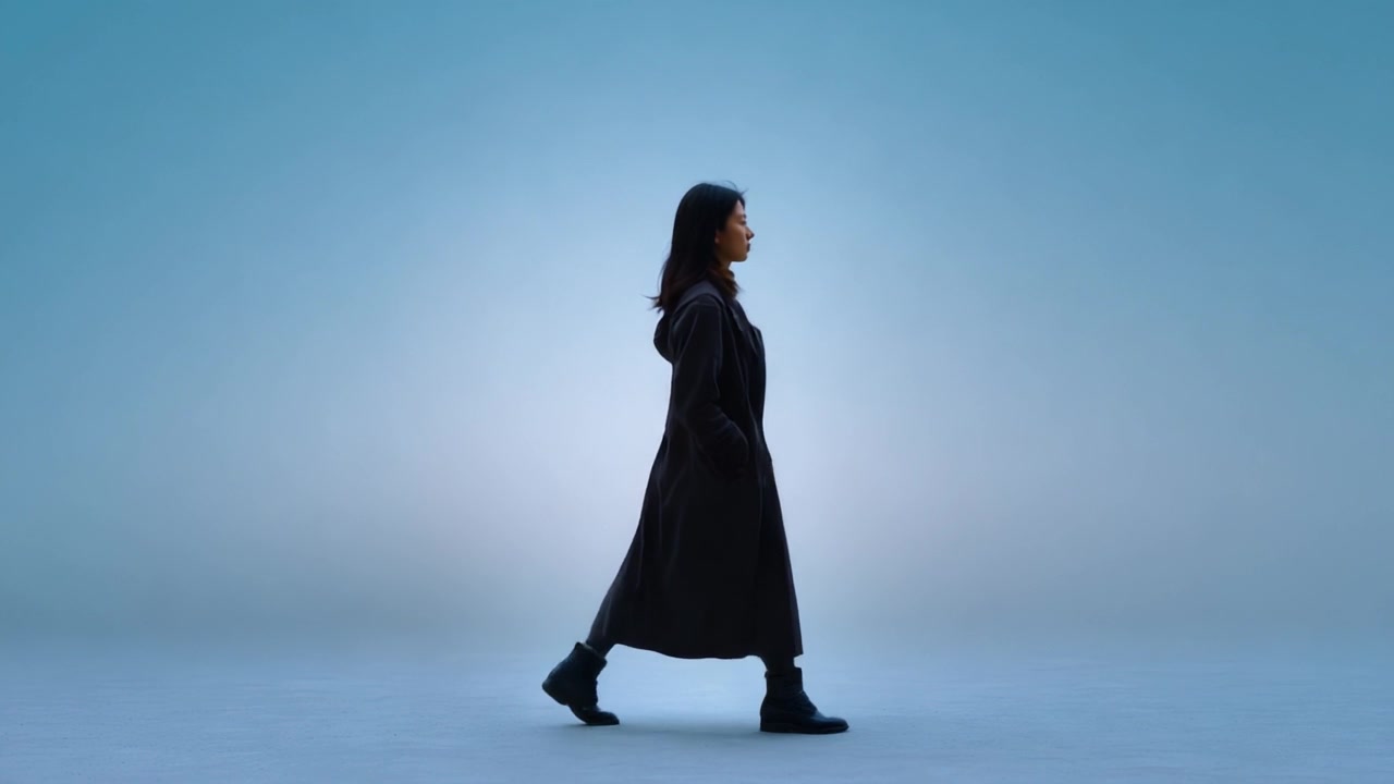 Una donna cammina con grazia contro uno sfondo blu sereno, incarnando eleganza e sicurezza nel suo mantello scuro e stivali robusti, unendo compostezza e tranquillità.
