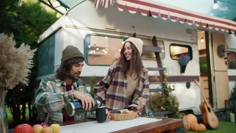 a happy brunette girl comes out of the trailer and brings out delicious pastries to her boyfriend while he pours tea and chats with his girlfriend while sitting at a table near the trailer outside the city during a picnic at the camp in the summer