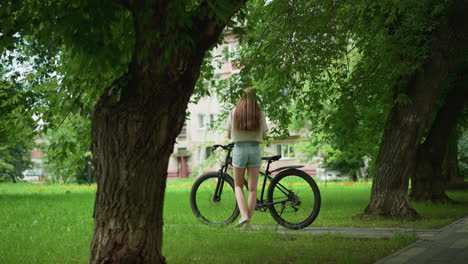 young woman in casual summer outfit approaches black bicycle parked on paved path, placing item on seat, background features lush greenery, towering trees, and residential building