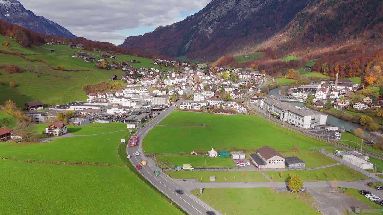 Traffic scene in Main Street between swiss villages during cloudy day in fall season. Colored trees on mountains. Green grass fields and cityscape in distance. Aerial wide shot