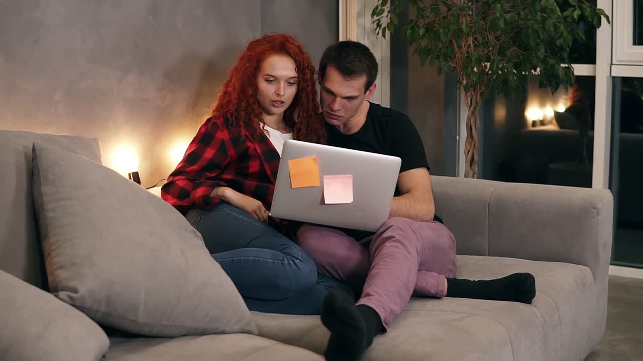 hermosa pareja de jóvenes sentados juntos con una computadora portátil gris en el sofá. pareja - hombre de cabello corto y chica de cabello rojo viendo algo en la computadora portátil sentado en el sofá en el elegante salón loft por la noche