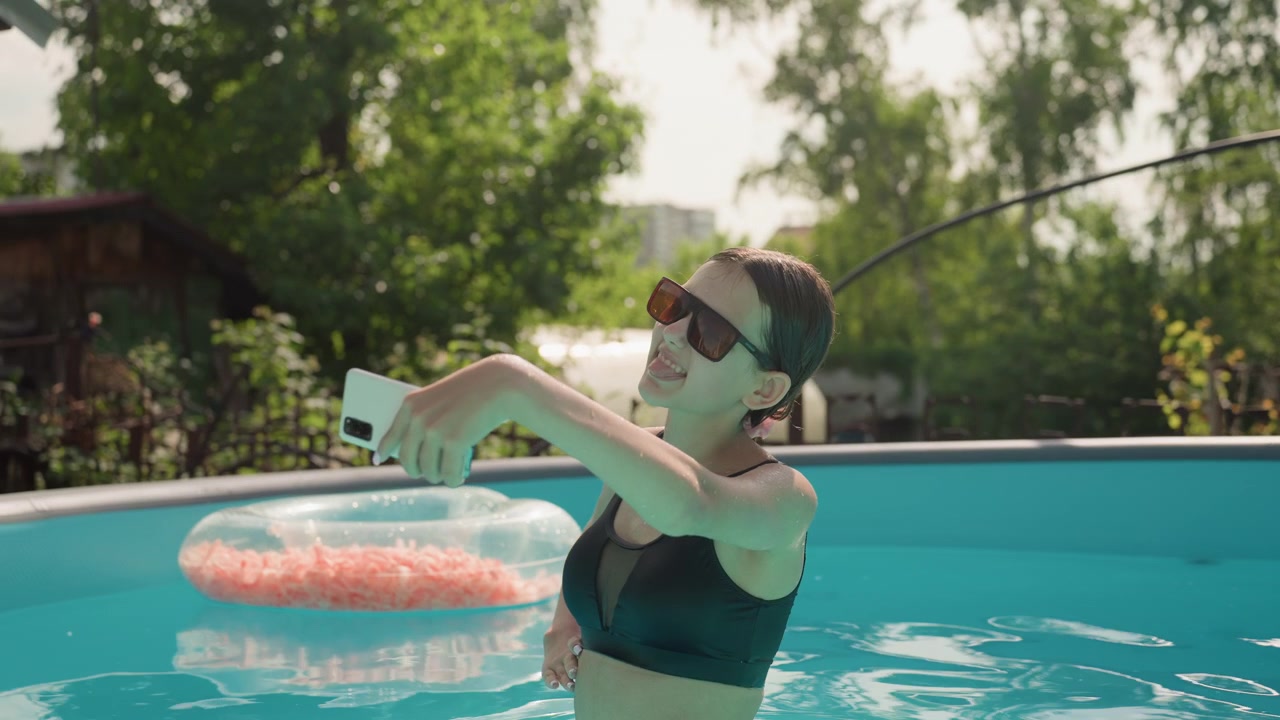 Woman in black swimsuit takes selfie with smartphone while standing waist deep in outdoor pool. Pool features clear blue water and pink inflatable ring set against green trees and bright