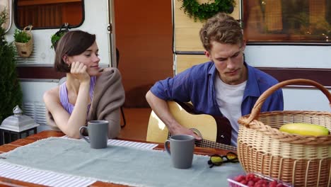 romantic couple sitting at the picnic table outdoors in front their home trailer and man playing the guitar for girlfriend. girl listening music passionately. slow motion