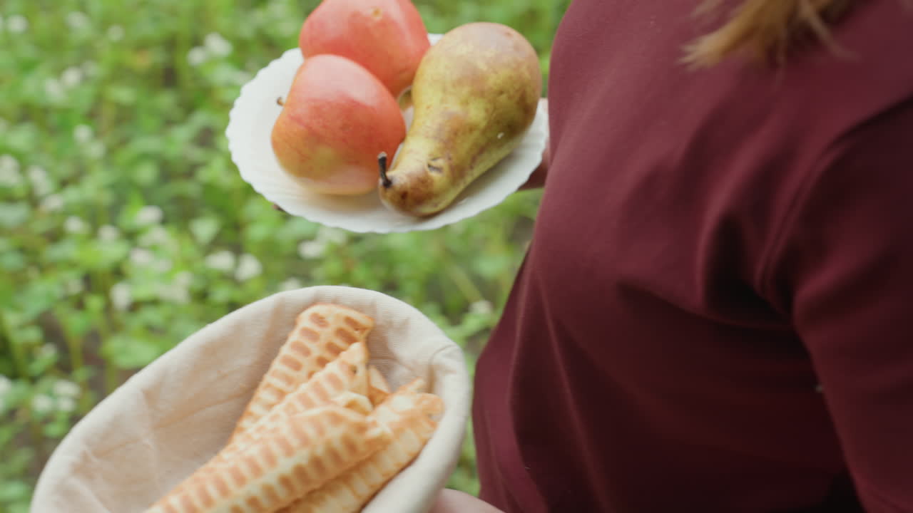 배와 바구니를 들고 녹색 뒤뜰을 가로지르는 백인 여성, 와플 콘이 담긴 천으로 안감을 댄 바구니와 사과 접시를 들고 있으며 데님 청바지와 샌들이 보이고 잔디와 흙이 있어요.