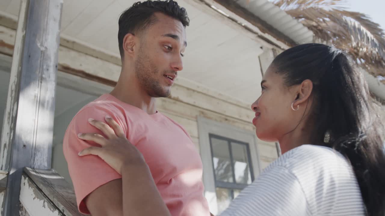 Happy diverse couple talking outside wooden beach house in the sun, in slow motion