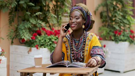 Mujer-Joven-Y-Guapa-Con-El-Traje-Tradicional-Hablando-Alegremente-Por-Teléfono-Mientras-Se-Sienta-En-La-Mesa-Con-Una-Revista-Y-Café-En-El-Hermoso-Patio