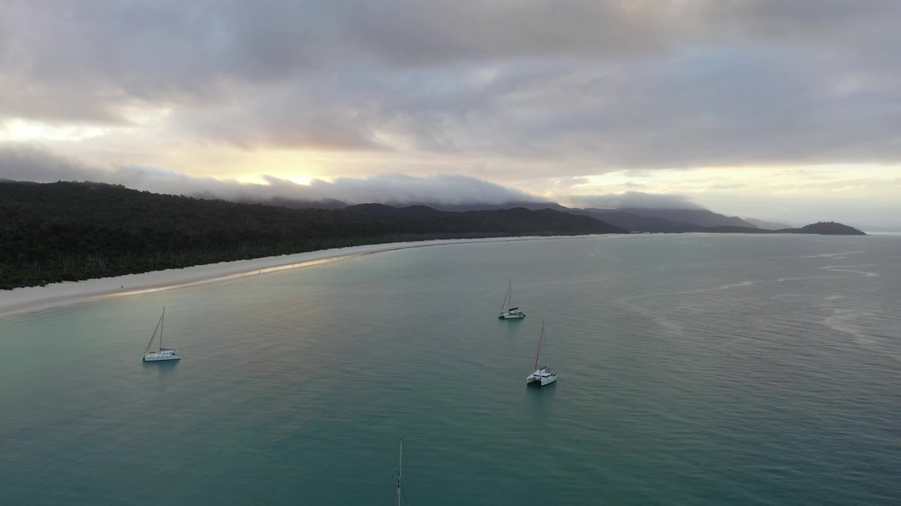 Vue aérienne du soir s'élevant au-dessus de catamarans à voile amarrés au large d'une plage de sable.