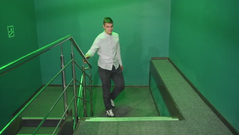 front shot of male in shirt and trousers walking up stairs, holding rail. serious expression under green lighting. shadows cast on walls, creating an atmospheric scene in an industrial-style interior