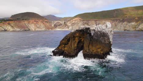 Vista-Aérea-De-Las-Olas-Del-Océano-Pacífico-Chocando-Contra-Una-Pequeña-Isla-Rocosa-Frente-A-Big-Sur