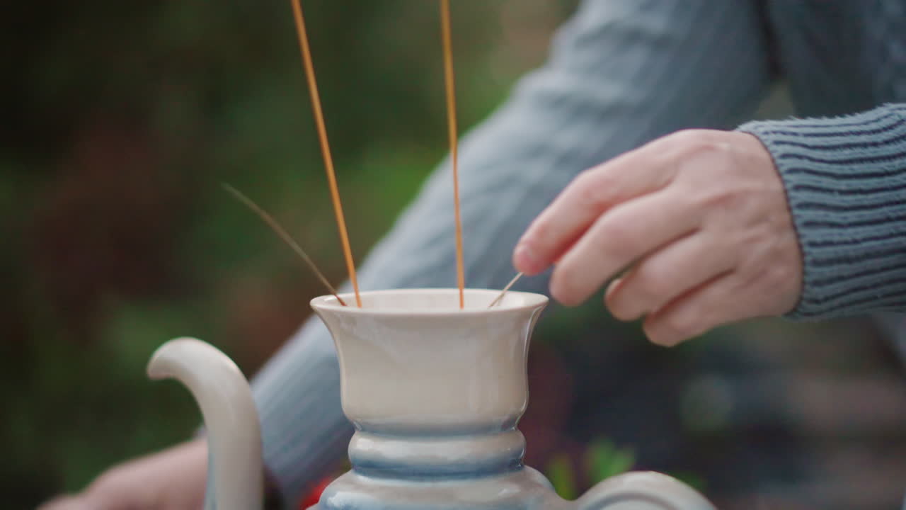 Primo piano di mani che sistemano steli secchi in una teiera blu, una stylist di interni sistema le erbe in un vaso di ceramica su un tavolo coperto di pizzo, maniche di un maglione accogliente, movimento lento e attento, preparazione di un centrotavola rustico.