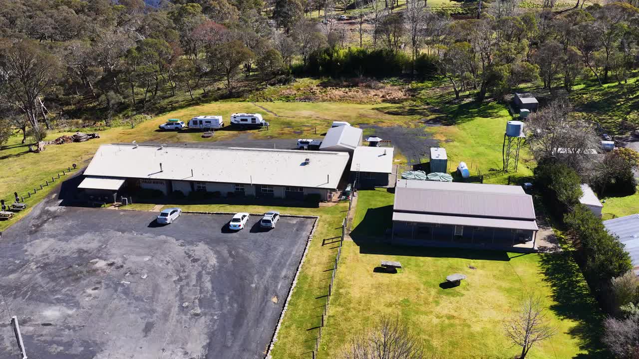 Drone camera moves steadily toward a rural house with adjacent parking lot and outbuildings, under bright daylight in a green, wooded Australian landscape