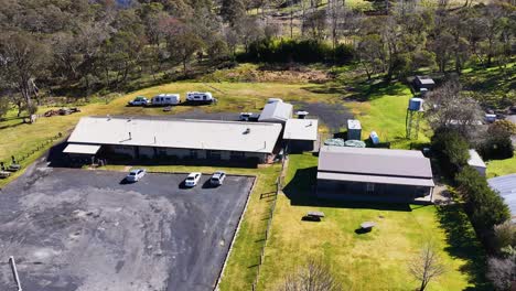 drone camera moves steadily toward a rural house with adjacent parking lot and outbuildings, under bright daylight in a green, wooded australian landscape