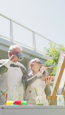 vertical video of happy diverse senior couple painting in garden on sunny day