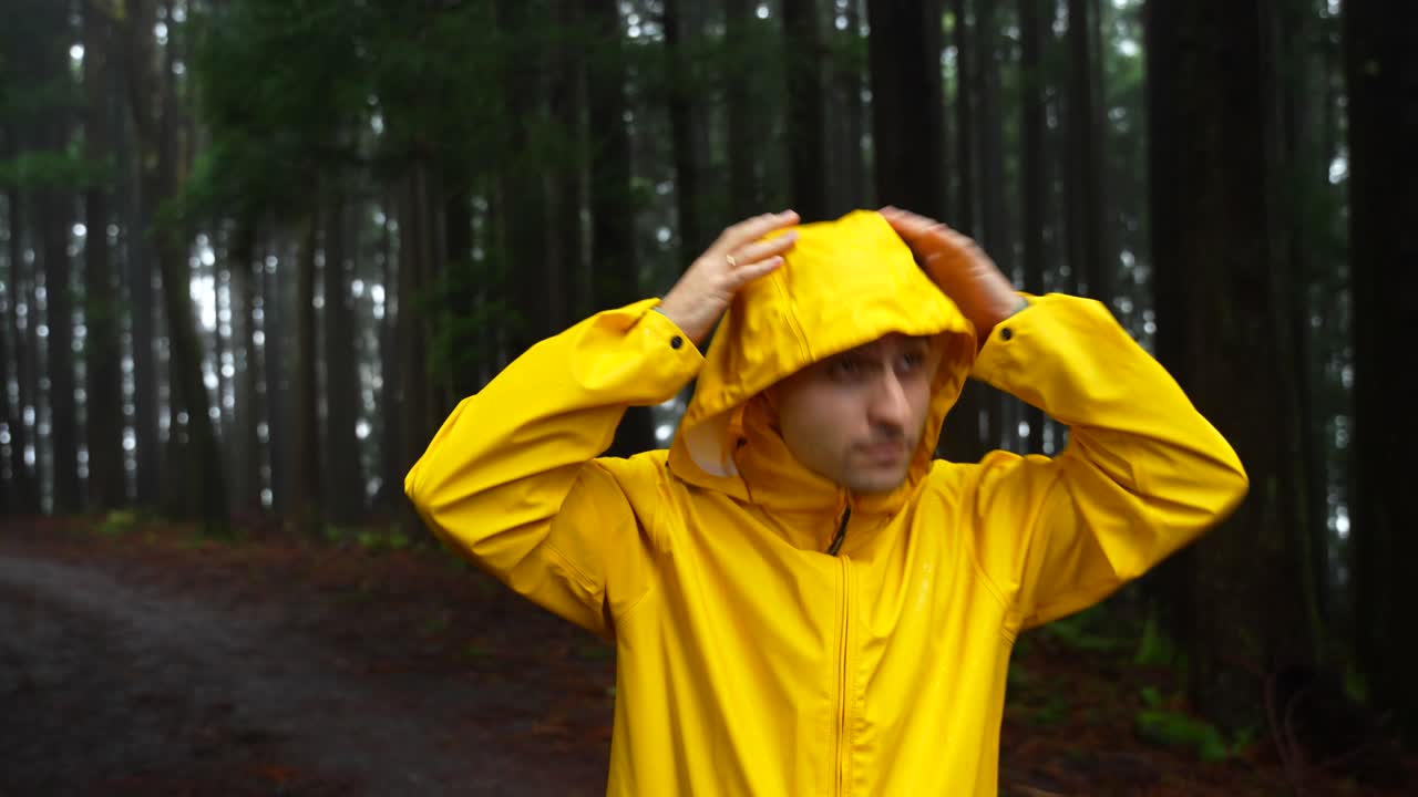 Young man zipping bright yellow raincoat in dark rainy forest, looking around