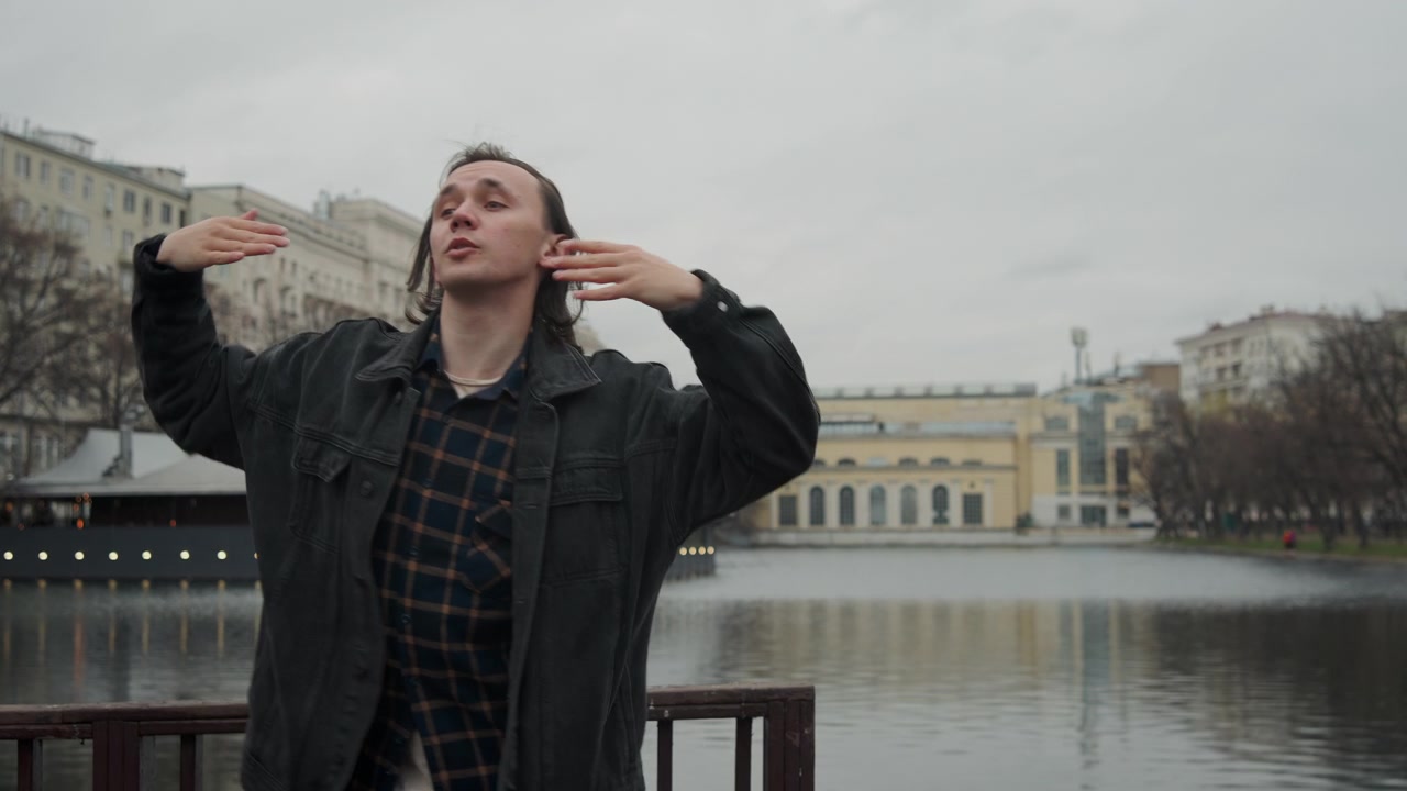 Мужчина с длинными волосами выполняет выразительные жесты и движения руками на мосту над водой, одетый в темную куртку. На заднем плане — классическая архитектура, голые деревья, белые палатки.