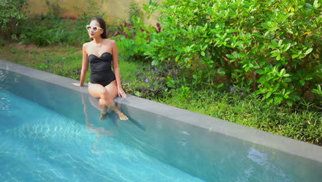 a pretty young woman in a black one-piece bathing suit sits on the side of a swimming pool with her feet in the water