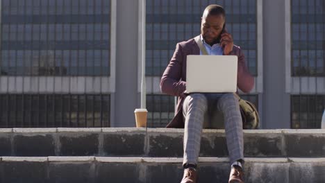 african american man in city sitting on stairs with coffee, using smartphone and laptop