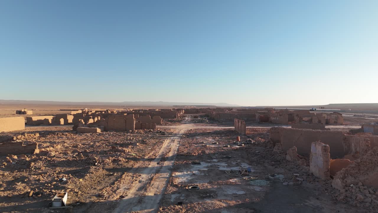 Drone shot moving forward above abandoned nitrate town ruins in the Atacama Desert, Chile. Crumbling walls, debris and vast arid landscape under a clear blue sky at golden hour.