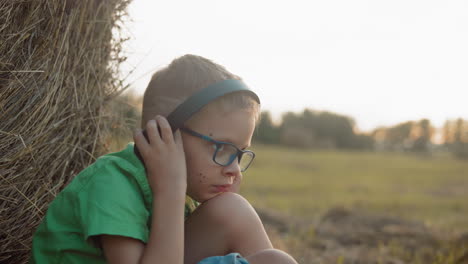 side view of a little child seated, adjusting his headphone, resting against a hay bale with spots on his cheek in a serene countryside setting