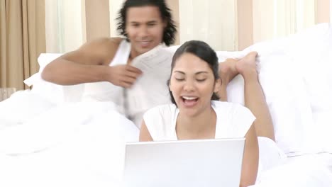 woman using a laptop and man reading a newspaper in bed