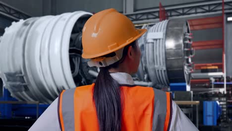 close up back view of a female engineer wearing safety helmet looking around while standing with arms akimbo with airplane engine maintenance conducted