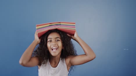 Happy-african-american-girl-holding-books-on-head-on-blue-background,-slow-motion