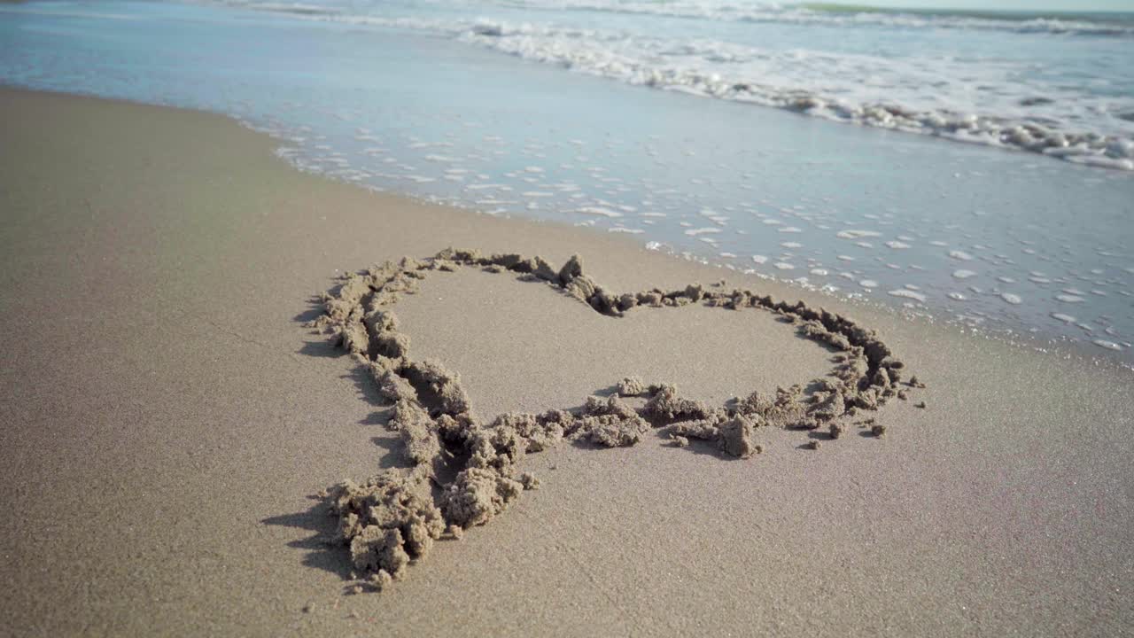 해변의 모래에 그린 하트 모양이 파도에 의해 부분적으로 씻겨 나가고 있습니다.