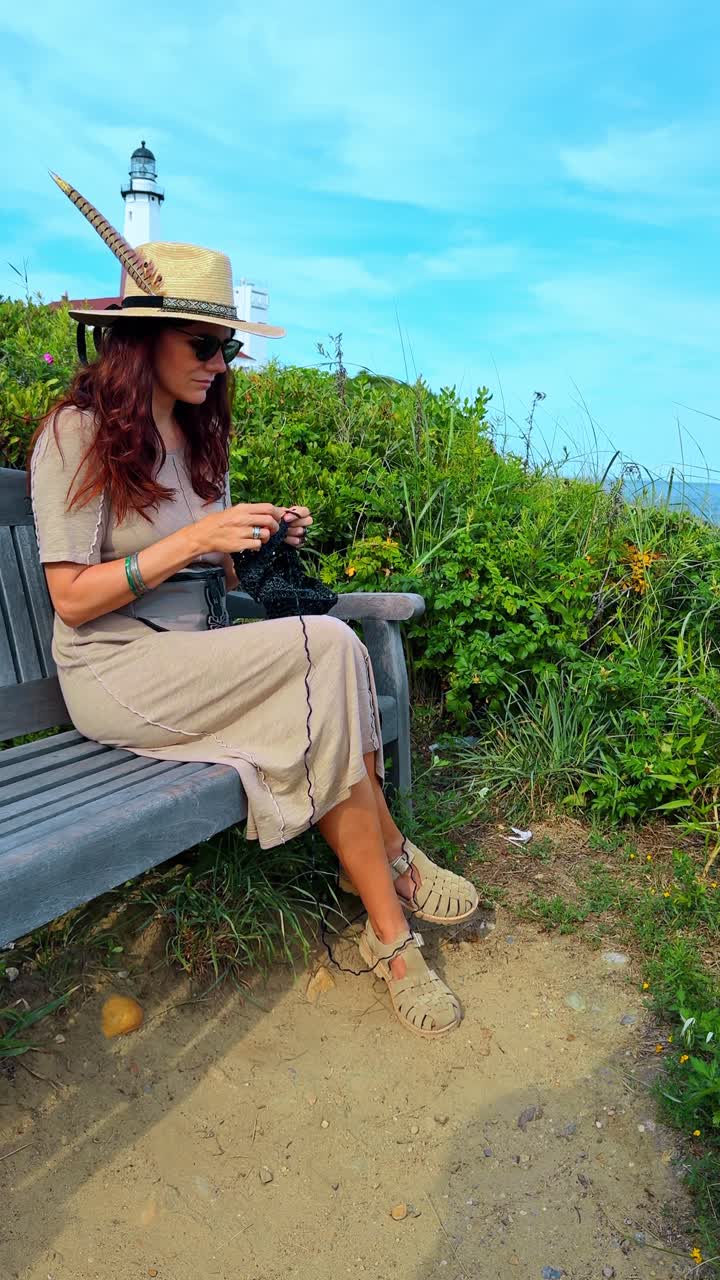 Viaggiatrice con cappello da sole che lavora a maglia su una panchina in un parco costiero. Donna in un abito lungo e cappello di paglia concentrata sul progetto di maglia seduta su una panchina di legno in riva al mare