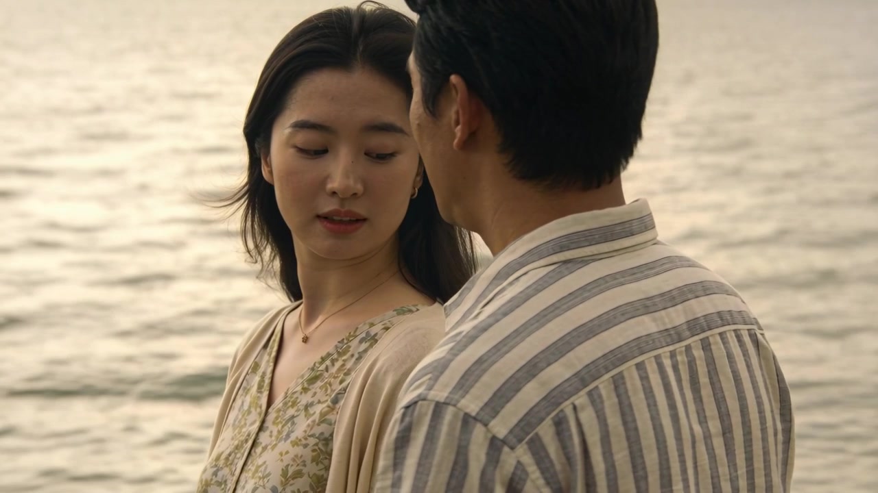 岸辺で、男性の方を向き体を傾ける女性と、視線を返す寄り添うパートナー。女性はペンダントとストライプの服を着用。