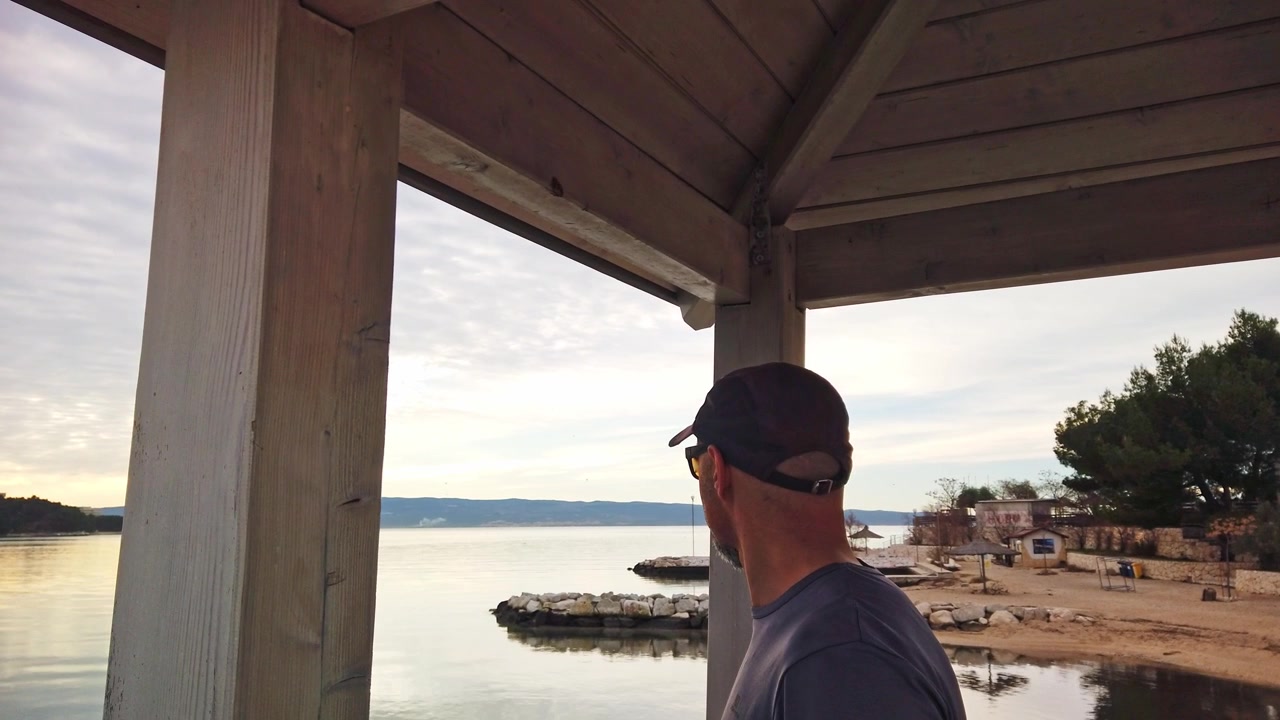 Side view of man with cap looking at calm sea bay from wooden gazebo structure. Concept for coastal surveillance, vacation break, travel destination, and quiet moment.