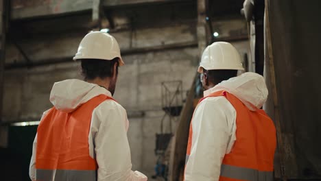 a man with a beard in a white helmet and a white protective uniform stands and communicates with his colleague a man with black skin in an orange vest near a conveyor belt at a large gray waste recycling plant