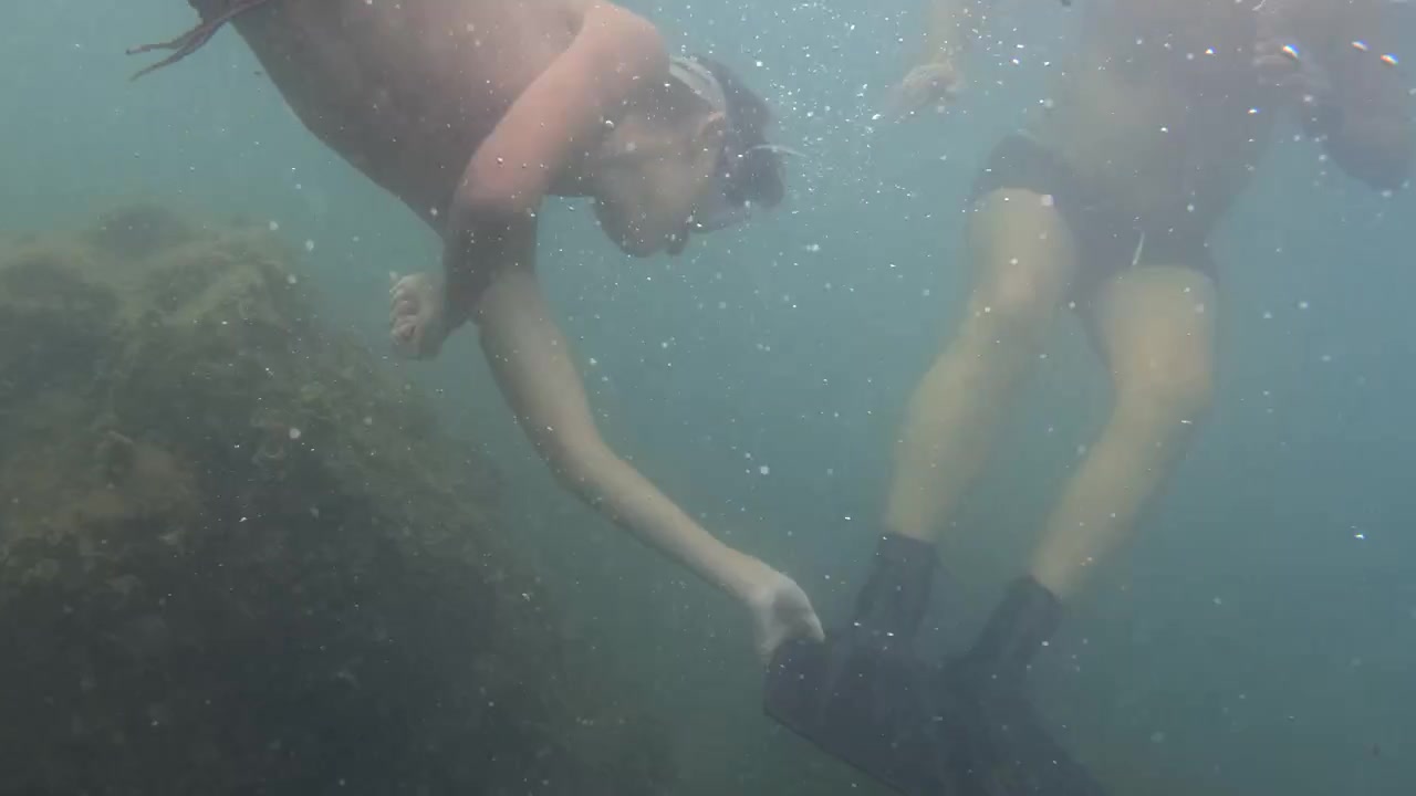 Mand med snorkelmaske svømmer under vandet og udforsker havbunden. Bobler stiger op gennem det blågrønne vand.