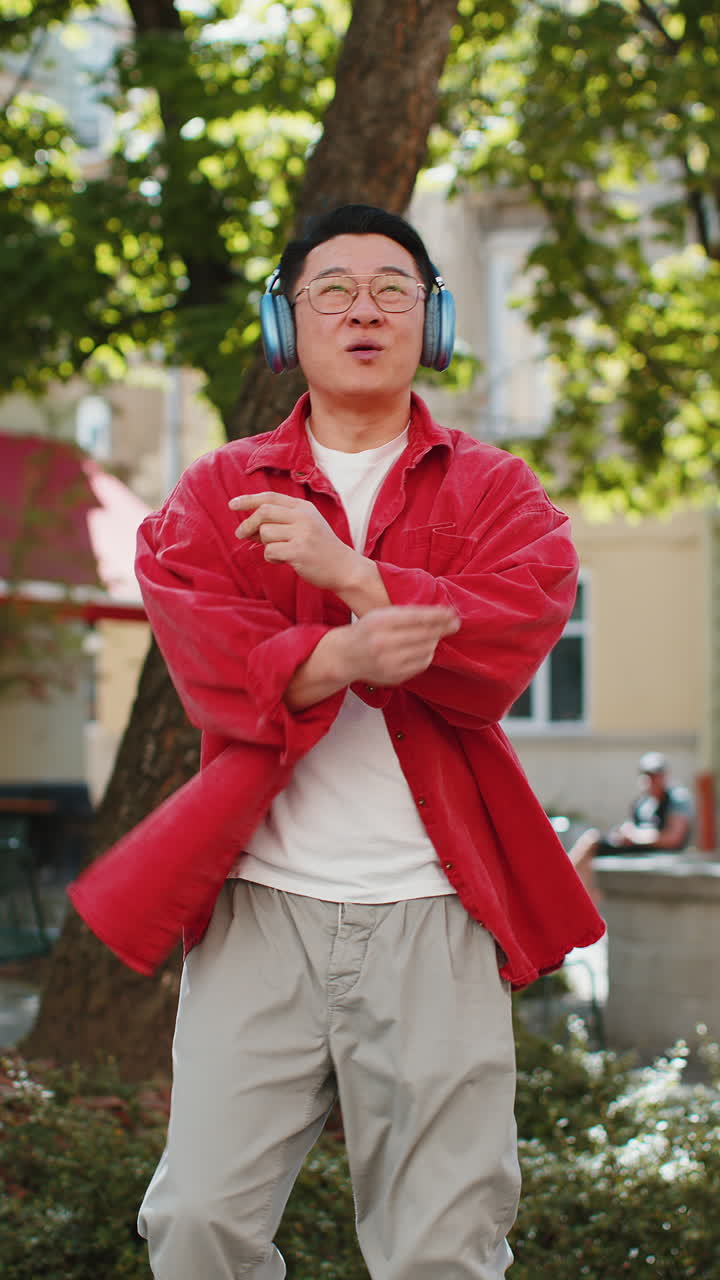 Happy asian man dancing in wireless headphones listening favorite energetic music on city street