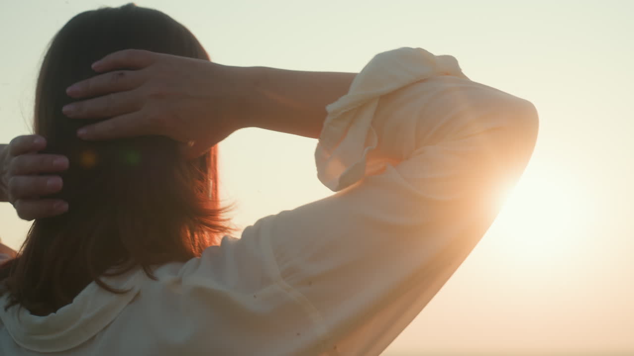 Rear view of young woman in white shirt raising arms to adjust hair while standing in open field during sunset, warm sunlight glowing around silhouette creating peaceful serene atmosphere