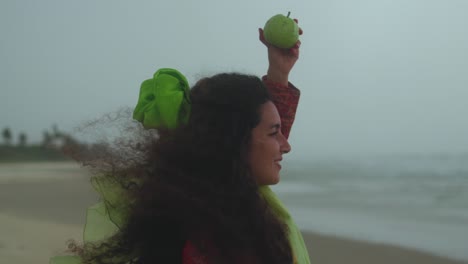 a smiling woman stands on the beach, holding a green apple above her head. fixed shot. she wears a red dupatta, green sari, and has curly hair with a green hair tie