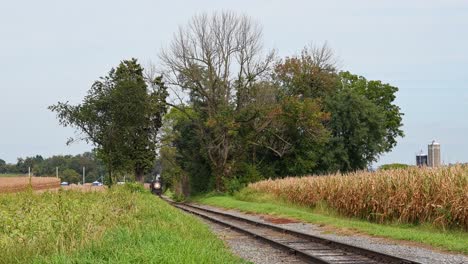 una vista de un antiguo tren de pasajeros de vapor restaurado que se acerca a través de un corte en los campos de maíz, soplando humo y vapor en un día soleado